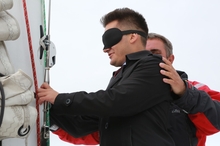Мужчина в черной ветровке и маске для сна тактильно осматривает такелаж яхты. Его придерживает за плечо Олег Колпащиков в красно-черной куртке. 
